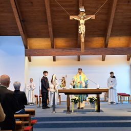 Ein Priester steht am Altar, umgeben von Gehilfen mit Kerzen, in einer Kirche mit einem großen Kruzifix darüber. Zuschauer sitzen auf den Kirchenbänken.