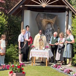 Ein Priester führt eine Zeremonie in einer Freiluftkapelle durch, wobei Menschen um einen geschmückten Tisch stehen und eine Statue im Hintergrund steht.