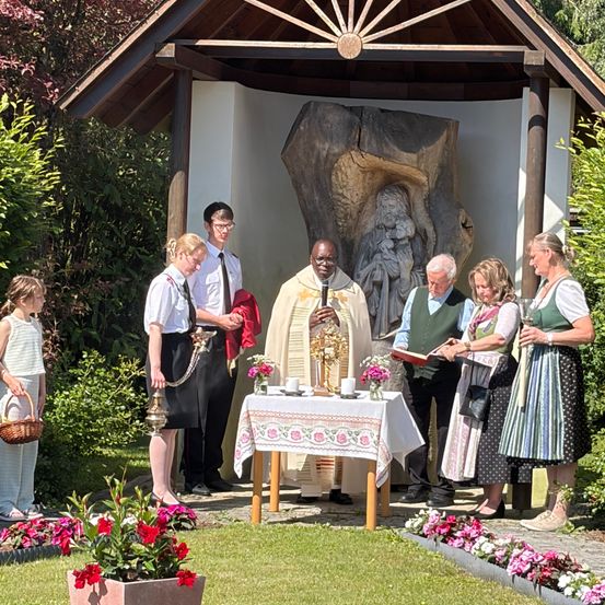 Ein Priester führt eine Zeremonie in einer Freiluftkapelle durch, wobei Menschen um einen geschmückten Tisch stehen und eine Statue im Hintergrund steht.