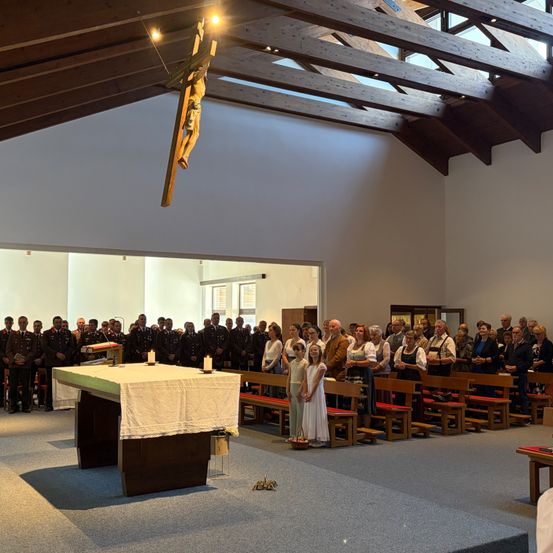 Eine Kirchenmesse ist im Gange mit einer großen Gemeinde, einem Altar und einem Kruzifix, das an der Decke hängt. Menschen stehen vor dem Altar, während andere auf hölzernen Bänken sitzen.