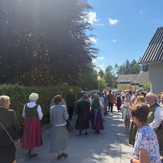 Eine Menschenmenge in traditioneller Kleidung geht auf einem Betonweg, mit Bäumen und Büschen auf beiden Seiten. Dahinter ist ein Gebäude mit einem schrägen Dach zu sehen.