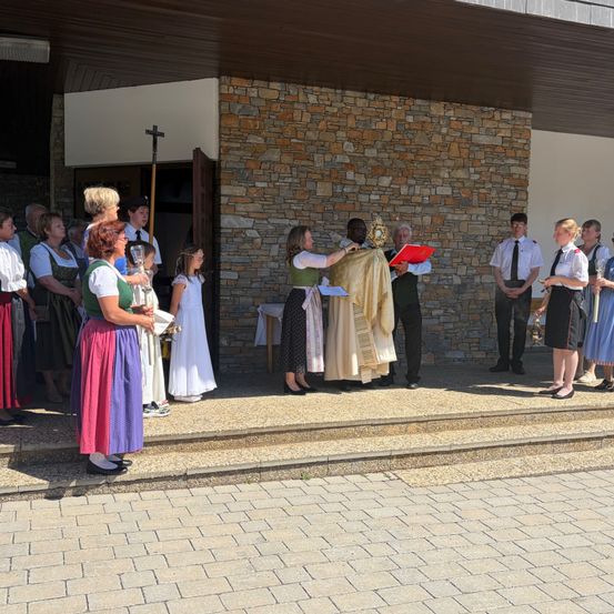 Ein Priester führt eine Zeremonie vor einer Kirche mit einer Gruppe von Menschen durch, darunter ein Mädchen mit einer Kerze.