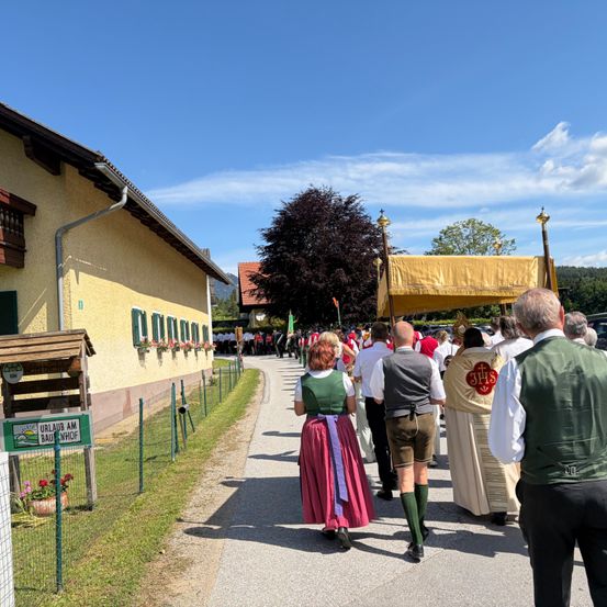 Menschen in traditioneller Kleidung gehen während eines Umzugs eine Straße entlang. Ein Haus mit einem Schild, das 'Urlaub am Bauhof' besagt, befindet sich auf der linken Seite.