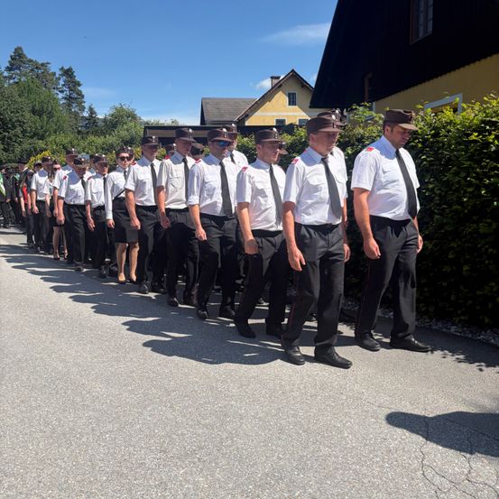 Eine Reihe uniformierter Personen, möglicherweise eine Marschkapelle, geht an einem sonnigen Tag die Straße entlang. Sie sind mit Bäumen und Gebäuden im Hintergrund ausgerichtet.