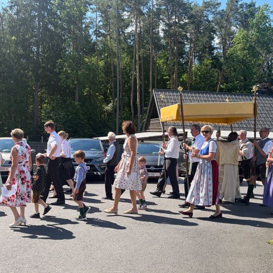 Eine Gruppe von Menschen in traditionellen Kostümen, die in einem Waldgebiet in einer Prozession gehen, mit einer überdachten Struktur im Vordergrund.