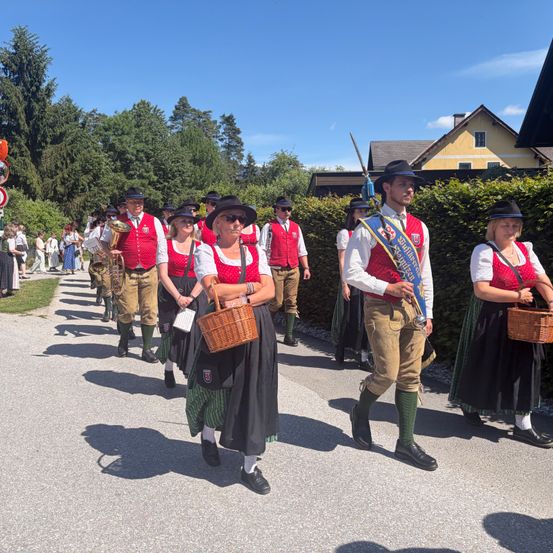 Eine Gruppe von Menschen in traditioneller bayerischer Tracht marschiert einen Pfad hinunter. Einige tragen Körbe und ein Mann hält eine Fahne.