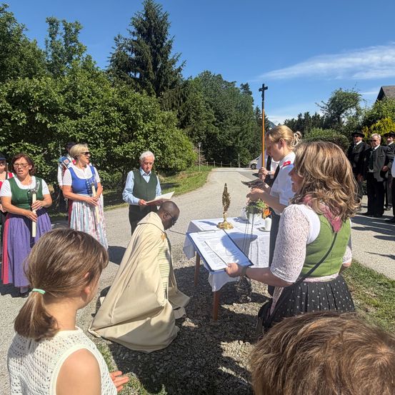 Eine religiöse Zeremonie an einem sonnigen Tag. Ein Priester kniet vor einem Tisch mit einem Kreuz und eine Frau steht neben ihm mit einem Papier. Mehrere Menschen in traditioneller Kleidung stehen in einem Kreis hinter ihnen.