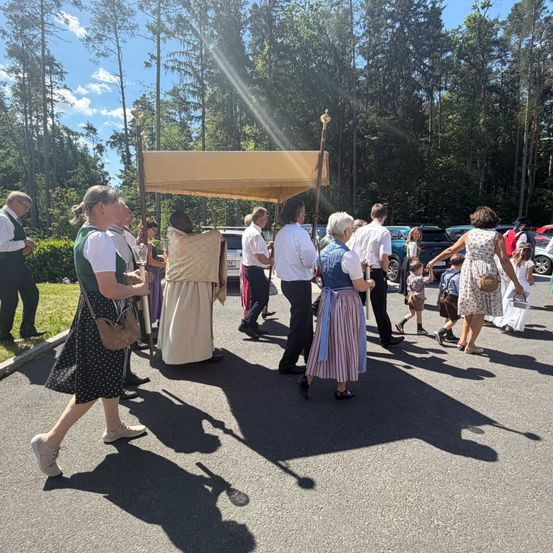 Eine Prozession von Menschen in traditioneller Kleidung geht unter einem Baldachin auf einem Parkplatz, mit Bäumen und geparkten Autos im Hintergrund.