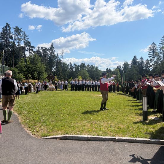 Eine Gruppe von Menschen in traditioneller Kleidung steht auf einer Rasenfläche, möglicherweise für ein Fest. Ein Mann in einem roten Vest führt eine Band an. Dahinter steht eine Menschenmenge und links steht ein Gebäude.