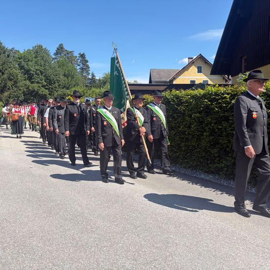 Eine Gruppe von Männern in formeller Kleidung, die an einem sonnigen Tag in einer Parade geht, mit Stäben und Flaggen.