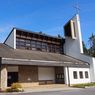 Eine moderne Kirche mit einem Kirchturm und einem Kreuz, umgeben von Bäumen und Pflanzen.