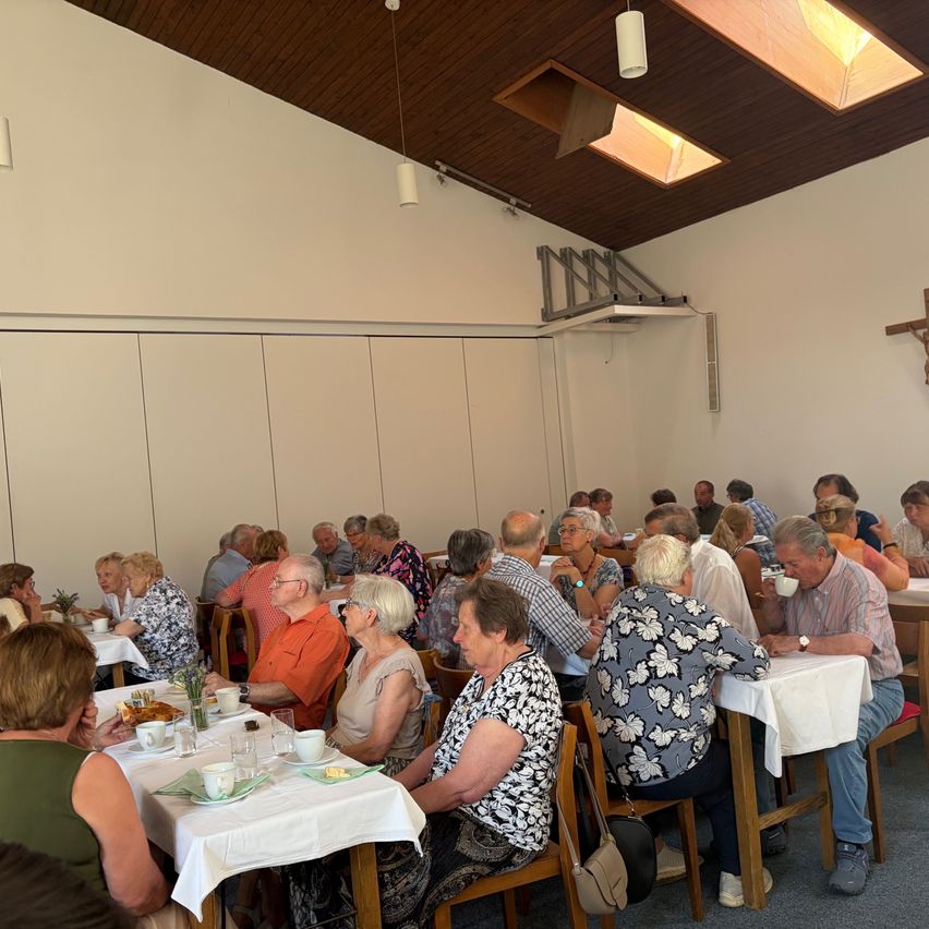 Eine große Gruppe älterer Menschen sitzt in einem Speisesaal um Tische herum. Sie unterhalten sich und essen. Der Saal hat eine Holzdecke mit Lichtern. Es gibt weiße Wände und weiße Tischdecken.