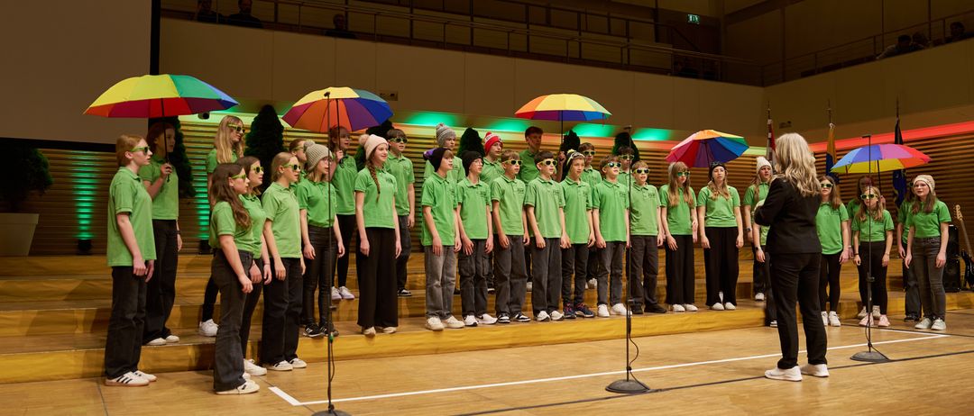Ein Chor von Kindern in grünen Hemden und schwarzen Hosen steht auf der Bühne, hält bunte Regenschirme und singt. Eine Frau steht rechts.