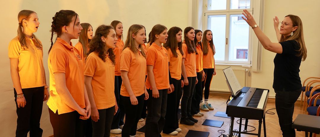 Ein Chor junger Frauen in orangefarbenen Hemden und schwarzen Hosen steht in einem Raum, singt, während ein Klavier vor ihnen steht und eine Person sie dirigiert.