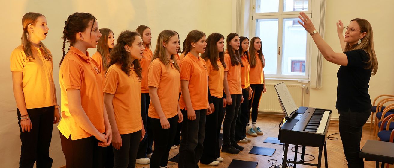 Ein Chor junger Frauen in orangefarbenen Hemden und schwarzen Hosen steht in einem Raum, singt, während ein Klavier vor ihnen steht und eine Person sie dirigiert.