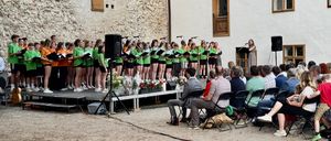 Ein Chor in grünen T-Shirts tritt vor einer Menge auf. Eine Frau singt an einem Podium mit Mikrofon. Das Publikum sitzt in Stühlen, und ein Mann spielt Gitarre. Die Bühne hat Blumen und Lautsprecher.