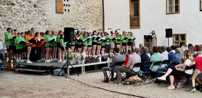 Ein Chor in grünen T-Shirts tritt vor einer Menge auf. Eine Frau singt an einem Podium mit Mikrofon. Das Publikum sitzt in Stühlen, und ein Mann spielt Gitarre. Die Bühne hat Blumen und Lautsprecher.