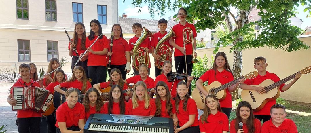 Ein Plakat für ein Schulkonzert zeigt eine Gruppe von Schülern in roten Hemden, die Instrumente spielen. Sie sind von Noten und Symbolen umgeben. Die Veranstaltung heißt 'Klassenkonzert der 2CM'.