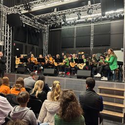 Eine Gruppe von Menschen spielt Gitarren und singt auf der Bühne in einem Studio. Das Publikum sitzt auf Stühlen. Die Bühne hat einen schwarzen Hintergrund und Metallkonstruktionen.