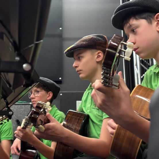 Drei junge Jungen in grünen Hemden und Mützen spielen Gitarren in einem Raum. Ein Junge ist im Vordergrund, ein anderer dahinter und ein dritter teilweise links sichtbar.