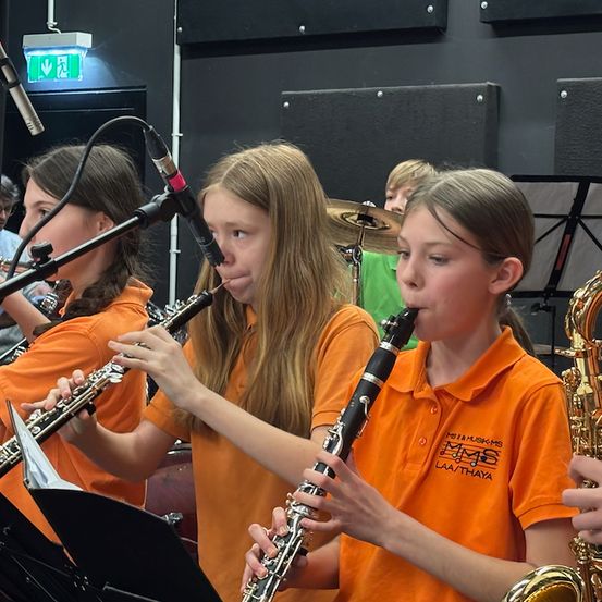 Mehrere Mädchen in orangefarbenen Uniformen spielen Blasinstrumente. Ein Mädchen vorne spielt Klarinette, ein anderes Flöte, und eine Posaune befindet sich rechts. Im Hintergrund sind weitere Schüler und Musikinstrumente zu sehen.