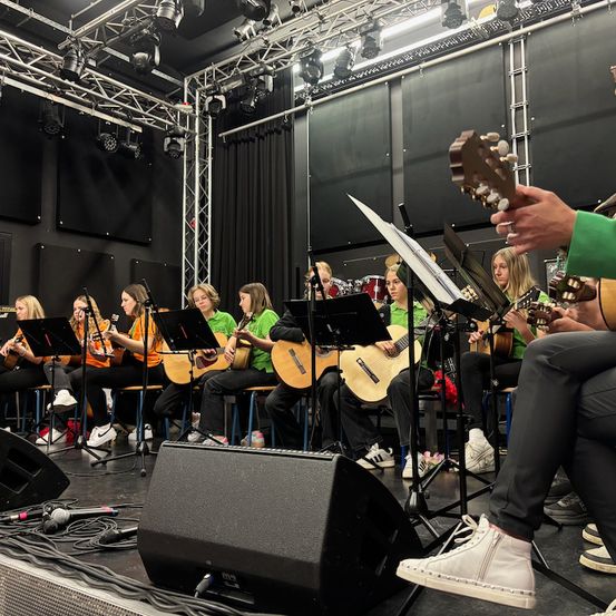 Eine Gruppe von Musikern in grünen Shirts spielt Gitarren auf der Bühne, mit Lautsprechern und Mikrofonen davor, unter Bühnenbeleuchtung.