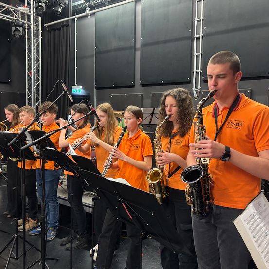 Eine Gruppe junger Musiker spielt in einem Studio mit schwarzen Wänden. Sie spielen verschiedene Instrumente, darunter Saxophone, Klarinetten und Flöten. Der Junge vorne spielt das Saxophon.