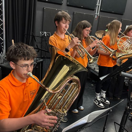 Eine Gruppe von Kindern in orangefarbenen Hemden und mit Brillen übt in einem Raum mit schwarzen Wänden und Vorhängen. Einige spielen Trompeten, während andere Tubas spielen.