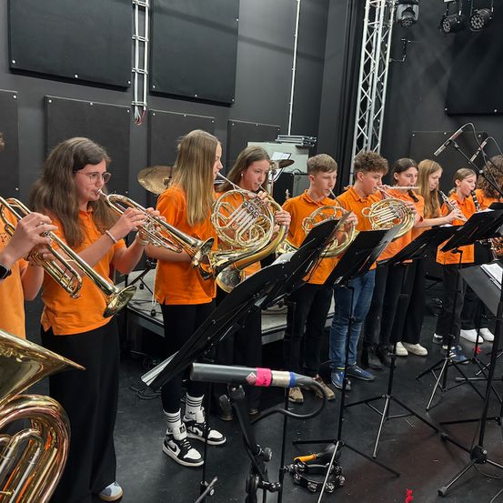 Eine Gruppe junger Musiker in orangefarbenen T-Shirts spielt in einem Studio ihre Instrumente. Sie halten Trompeten, Posaunen und Flöten, einige stehen, andere sitzen.