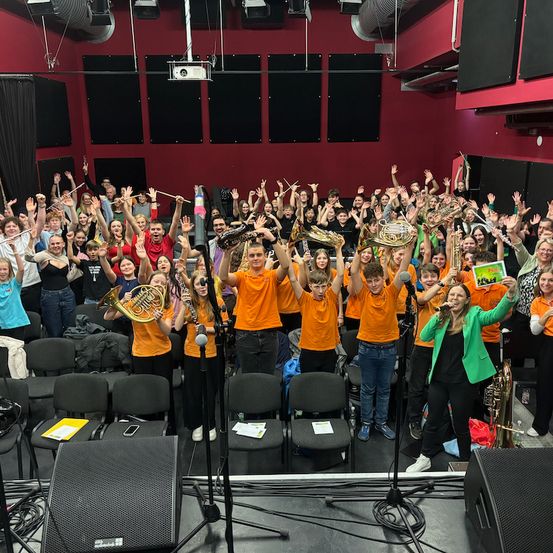 Eine große Gruppe von Menschen in orangefarbenen Shirts steht auf einer Bühne, hält Instrumente und hebt die Hände. Einige spielen Trompeten und Posaunen. Eine Frau in einem grünen Blazer steht rechts und singt in ein Mikrofon.