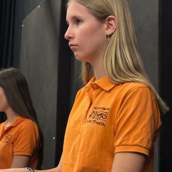 Zwei Frauen in orangefarbenen Shirts stehen nebeneinander, wahrscheinlich in einem Proberaum. Die Frau im Vordergrund hat lange blonde Haare und schaut nach links. Hinter ihr steht eine weitere Frau mit langen Haaren, die nach vorne schaut.