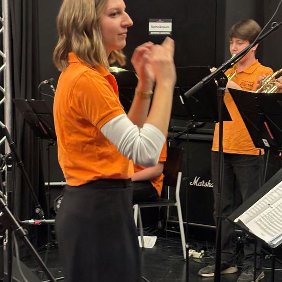 Eine Frau in einem orangefarbenen Shirt leitet eine Probe, während ein Junge hinter ihr Trompete spielt. Der Raum ist mit Musikinstrumenten und Ausrüstung gefüllt, was auf ein professionelles Umfeld hindeutet.