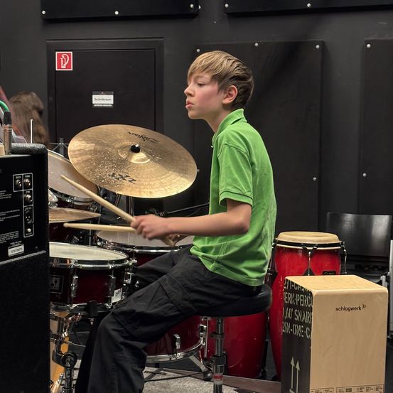 Ein junger Junge in einem grünen T-Shirt und schwarzen Hosen spielt die Trommeln in einem Musikraum. Er sitzt auf einem Hocker und hält Trommelstöcke. Hinter ihm sind verschiedene Trommeln und Becken angeordnet. Der Hintergrund ist eine schwarze Wand mit einem Schild.