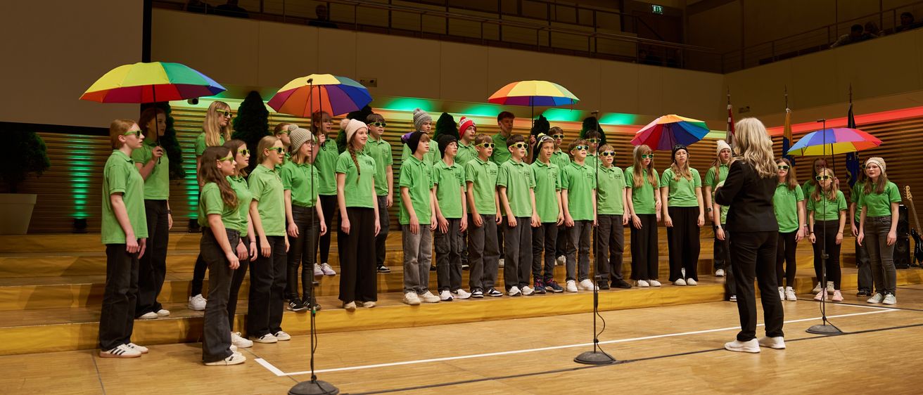 Ein Chor von Kindern in grünen Shirts steht auf der Bühne mit bunten Regenschirmen. Sie werden von einer Frau in Schwarz angeführt.