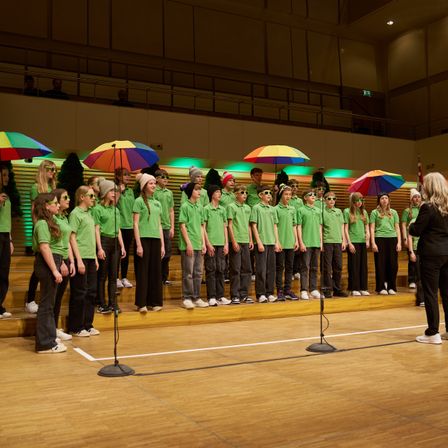 Ein Chor von Kindern in grünen Hemden tritt auf der Bühne auf, hält bunte Regenschirme in der Hand, eine Frau in Schwarz steht vor ihnen.