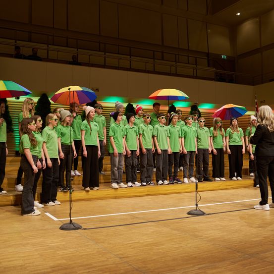 Ein Chor von Kindern in grünen Hemden tritt auf der Bühne auf, hält bunte Regenschirme in der Hand, eine Frau in Schwarz steht vor ihnen.