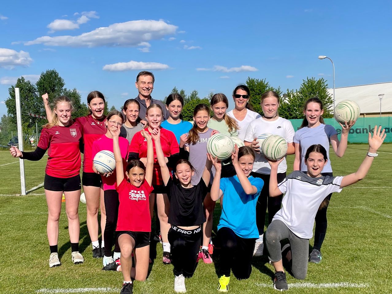 Eine Gruppe junger Mädchen und ein Mann posieren mit Rugbybällen auf einem Feld. Sie lächeln und tragen verschiedenfarbige Trikots. Bäume und eine Straßenlaterne sind im Hintergrund zu sehen.