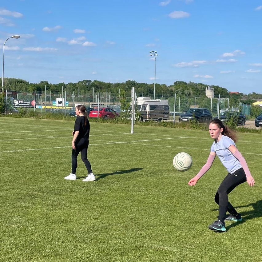 Zwei Mädchen spielen Fußball auf einem Rasenfeld, mit einem geparkten Auto und Wohnmobil im Hintergrund. Das Mädchen im Vordergrund bereitet sich darauf vor, den Ball zu treten.