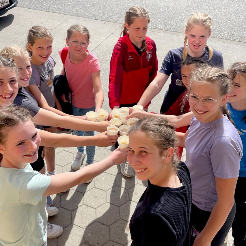 Eine Gruppe junger Mädchen versammelt sich draußen, hält Tassen mit einem Getränk in den Händen und lächelt in die Kamera. Der Boden ist mit sechseckigen Fliesen gepflastert.