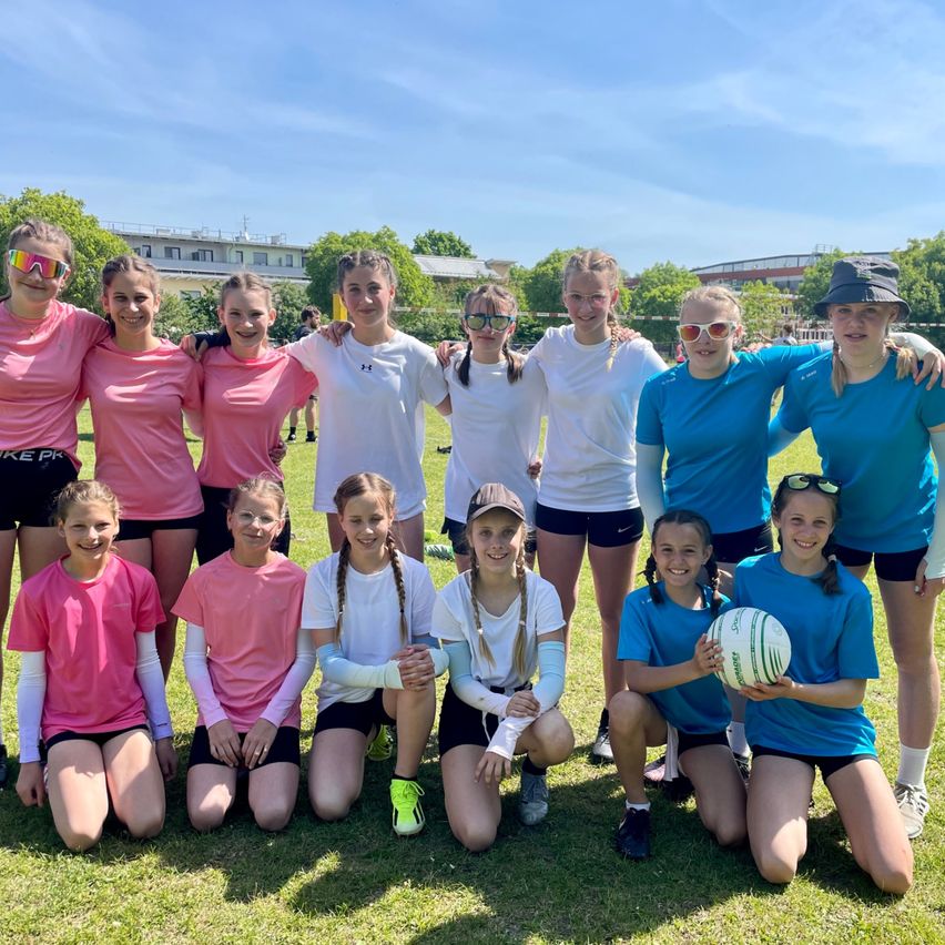Eine Gruppe junger Mädchen in pinken und blauen Sporttrikots posiert für ein Foto an einem sonnigen Tag in einem Park.
