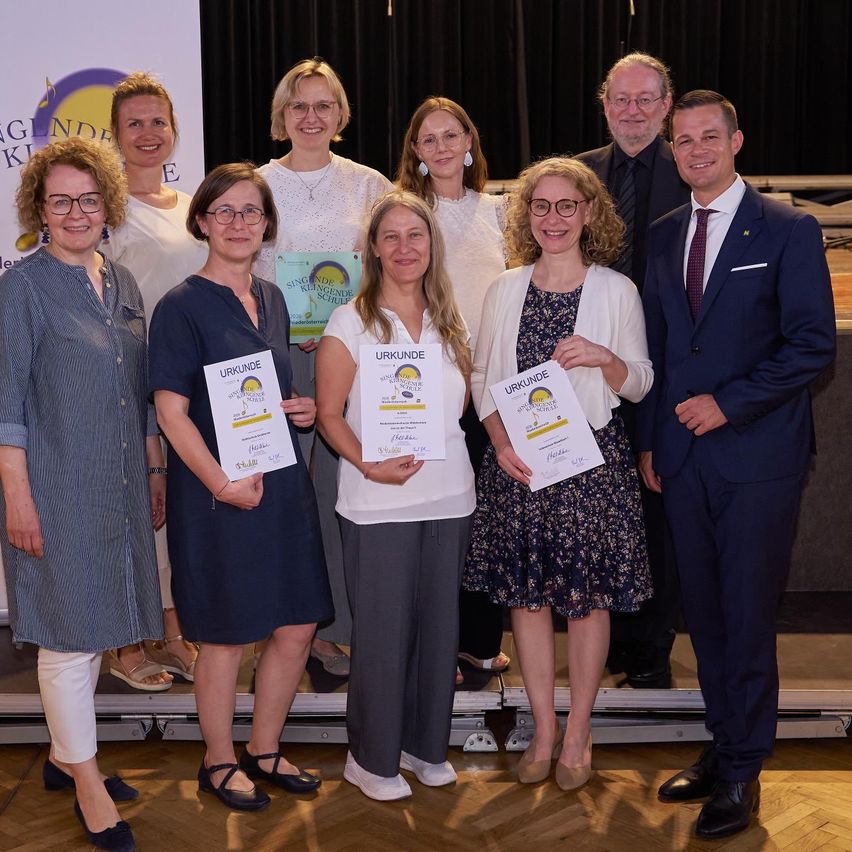 Eine Gruppe von Menschen, darunter Frauen und Männer, stehen lächelnd auf einer Bühne und halten Urkunden in der Hand. Sie sind in formeller Kleidung gekleidet. Hinter ihnen befindet sich ein Hintergrund mit Text.