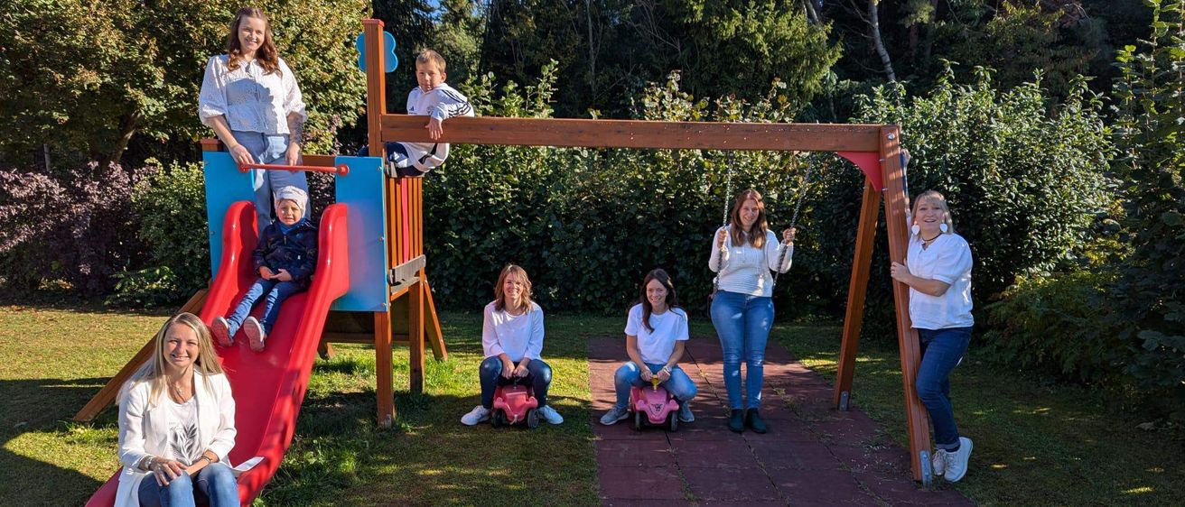 Eine Gruppe von Frauen und Kindern posiert auf einem Spielplatz mit Rutsche und Schaukeln. Ein Kind klettert auf die Rutsche, während andere auf den Schaukeln sitzen.