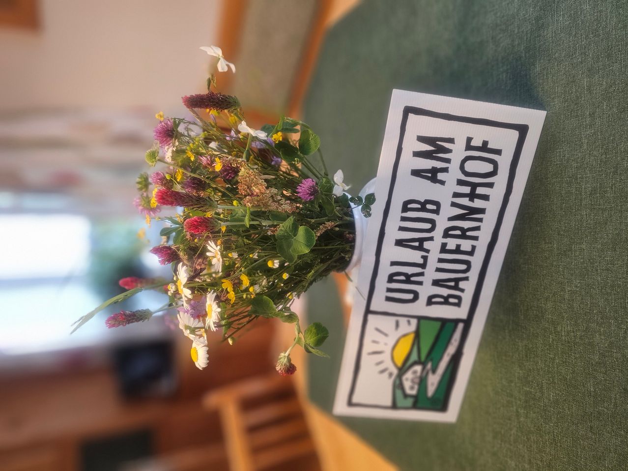 A white vase with a bouquet of wildflowers sits on a green table with a sign that says Urlaub am Bauernhof.