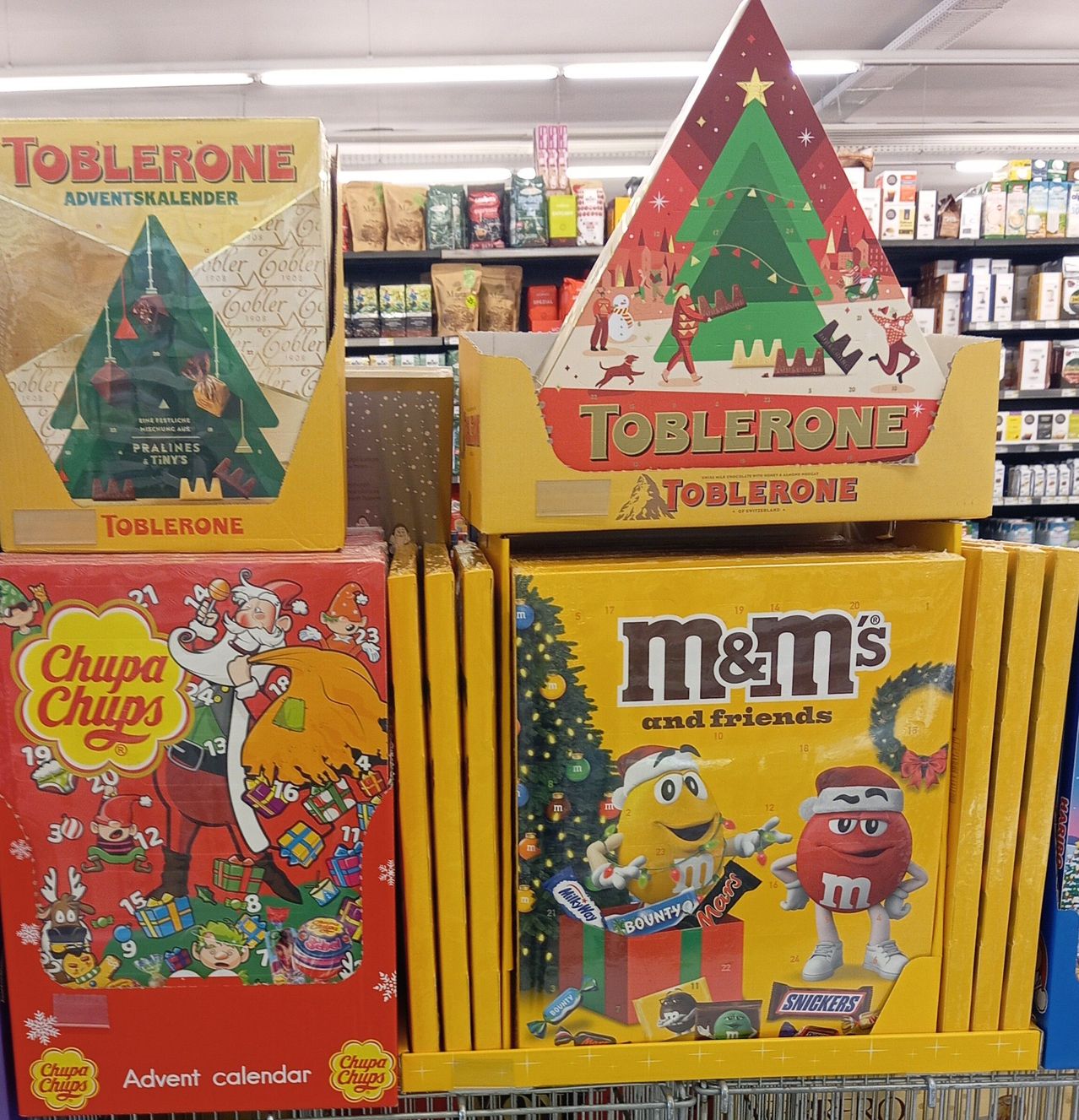 Ein Geschäft zeigt festliche Adventskalender mit Toblerone- und M&M's-Themen. Toblerone ist in einer dreieckigen Box mit einem Weihnachtsbaumdesign, während M&M's einen Geschenkkasten mit einem Weihnachtsbaum und Charakteren zeigt.