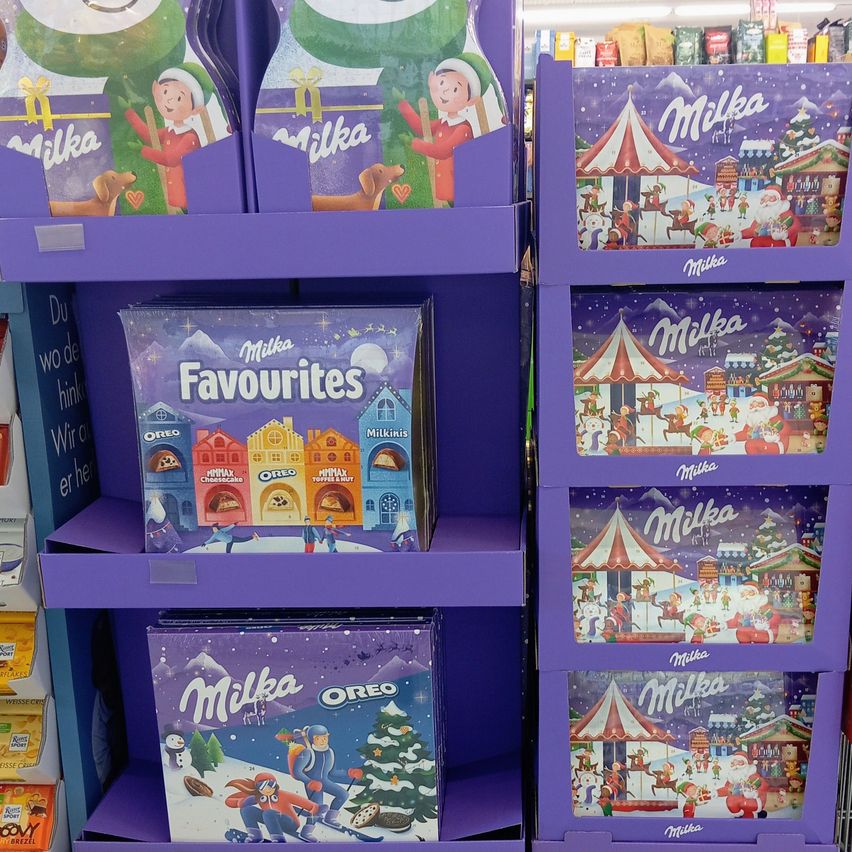Ein Regal im Laden gefüllt mit verschiedenen Schachteln mit Schokoladen. Die Schachteln sind lila und zeigen Bilder von Kindern, dem Weihnachtsmann und verschiedenen Feiertagsszenen. Eine Schachtel hat die Aufschrift 'Milka Favourites'.
