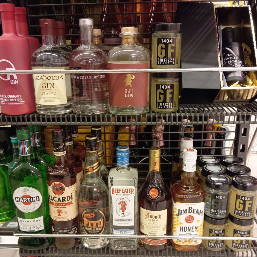 Ein Ladenregal zeigt verschiedene Likörflaschen, einschließlich Gin, Bacardi und Jim Beam, wobei einige eine rote Etikette und goldene Kappen haben.