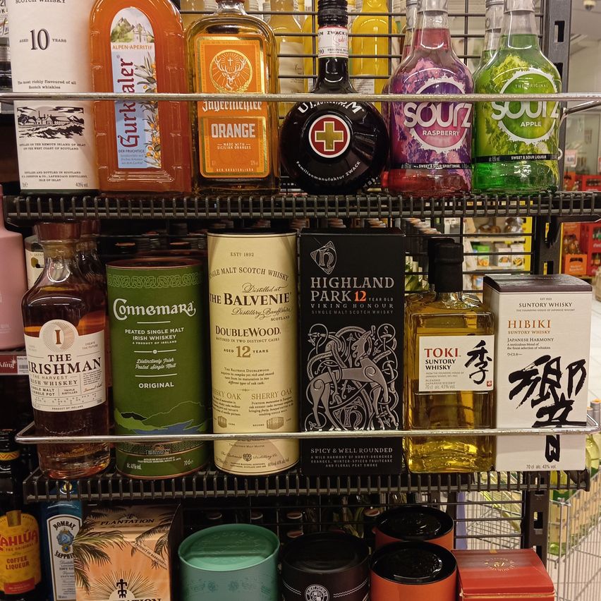 Ein Regal mit verschiedenen Alkoholmarken, darunter Scotch, Whisky und geschmackvolle Liköre. Im oberen Regal befinden sich Flaschen wie Gurkha, Orange und eine Marke mit rotem Kreuz. Darunter gibt es eine Vielzahl von Produkten wie Connemara, Dalvenvie, Highland Park, Toki und Hibiki.