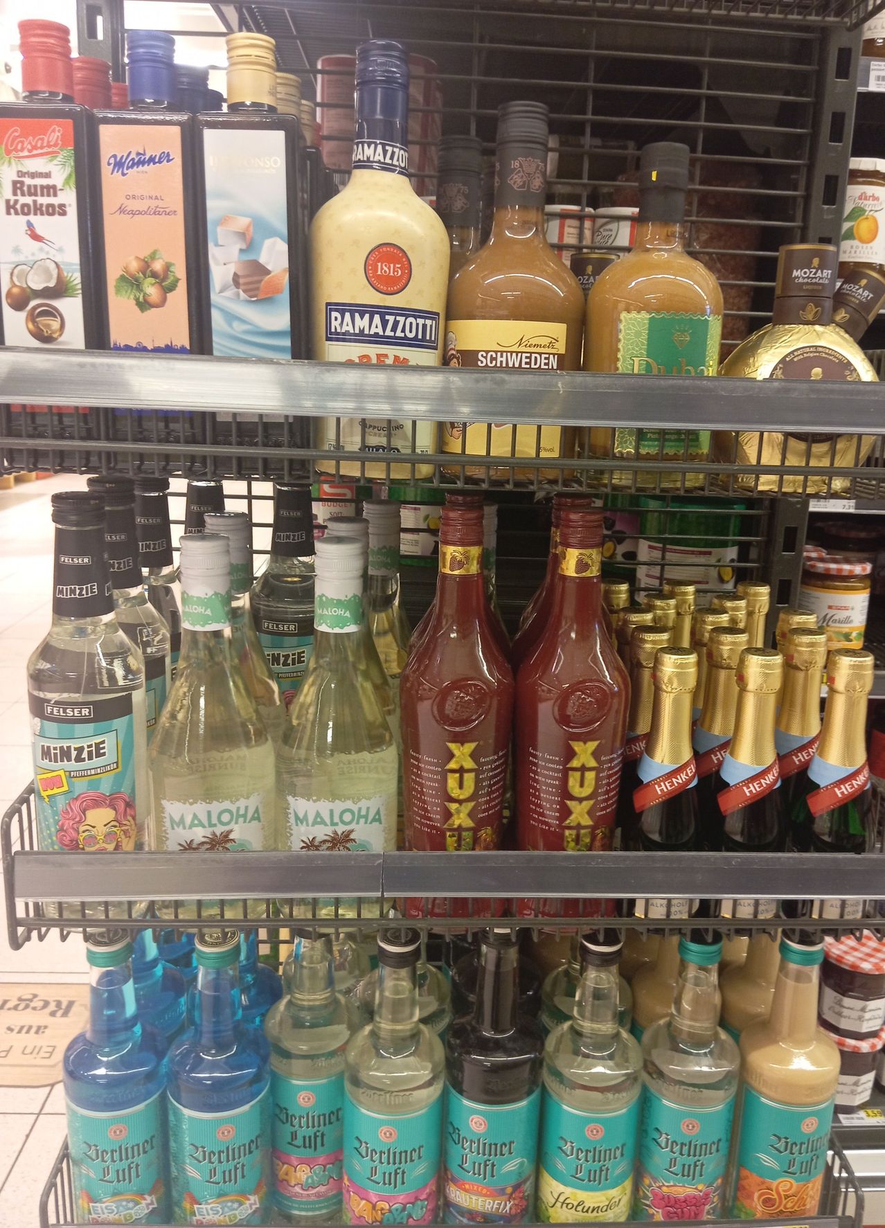 Ein Supermarktregal gefüllt mit verschiedenen Likören und Saucen. Die oberste Reihe hat Flaschen von Ramazotti, Nimet und Dubarry. Die mittlere Reihe hat Flaschen von XUX und die unterste Reihe hat Flaschen von Malhoa, Henckell und anderen Likören.