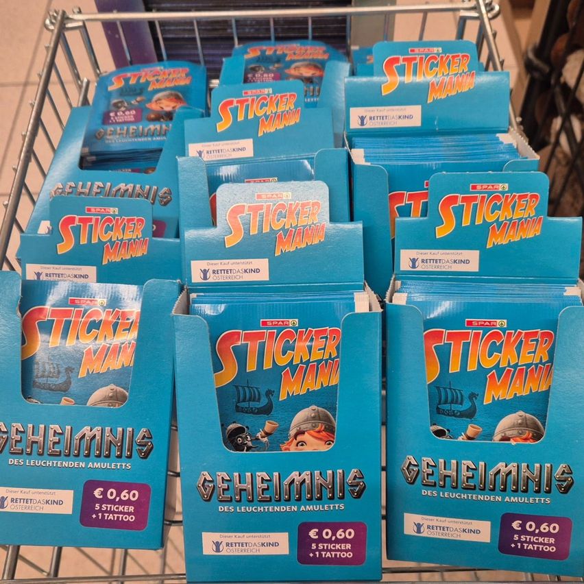 Ein blauer Einkaufswagen zeigt mehrere Packungen von Sticker Mania, einem Kinderstickerset, zum Preis von 0,60 €. Jede Packung enthält 5 Sticker und ein Tattoo. Der Hintergrund ist eine schlichte weiße Wand.