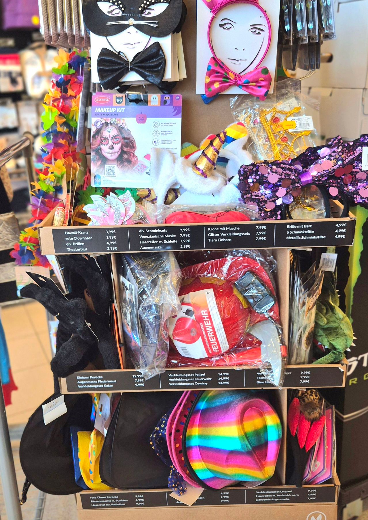 Eine Ausstellung verschiedener Kostüme und Accessoires in einem Geschäft. Es gibt Artikel wie Hawaii-Kranz, Gesichtsmasken, Bögen und Perücken. Ein Schild mit Preisen und Artikeln ist ebenfalls sichtbar.
